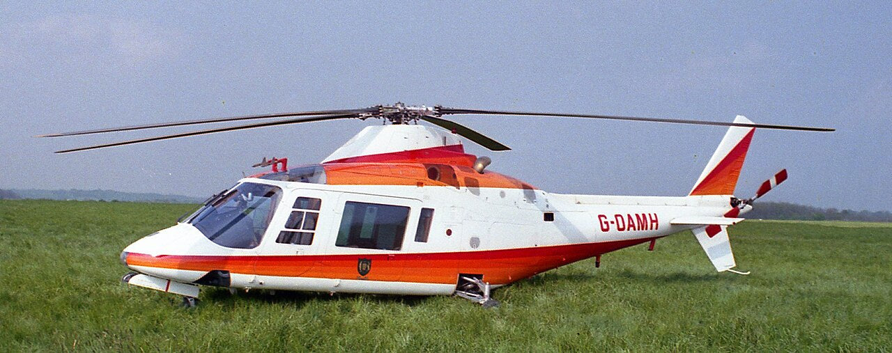 Red-and-white Agusta A109A G-OAMH parked in grass, with the full main rotor, nose and tail inside the frame; grass obscures the landing gear.