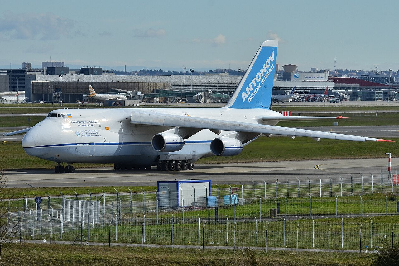 White-and-blue Antonov An-124-100 UR-82029 at Toulouse-Blagnac, seen from the front left with its high wings, large cargo fuselage and many landing wheels visible.
