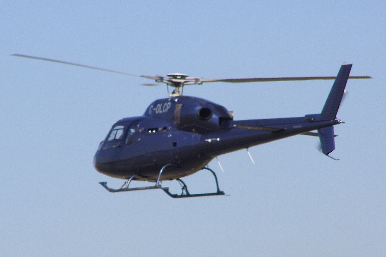 Blue Eurocopter AS355N G-OLCP in flight, with the fuselage, tail, rotor span and skids in view.