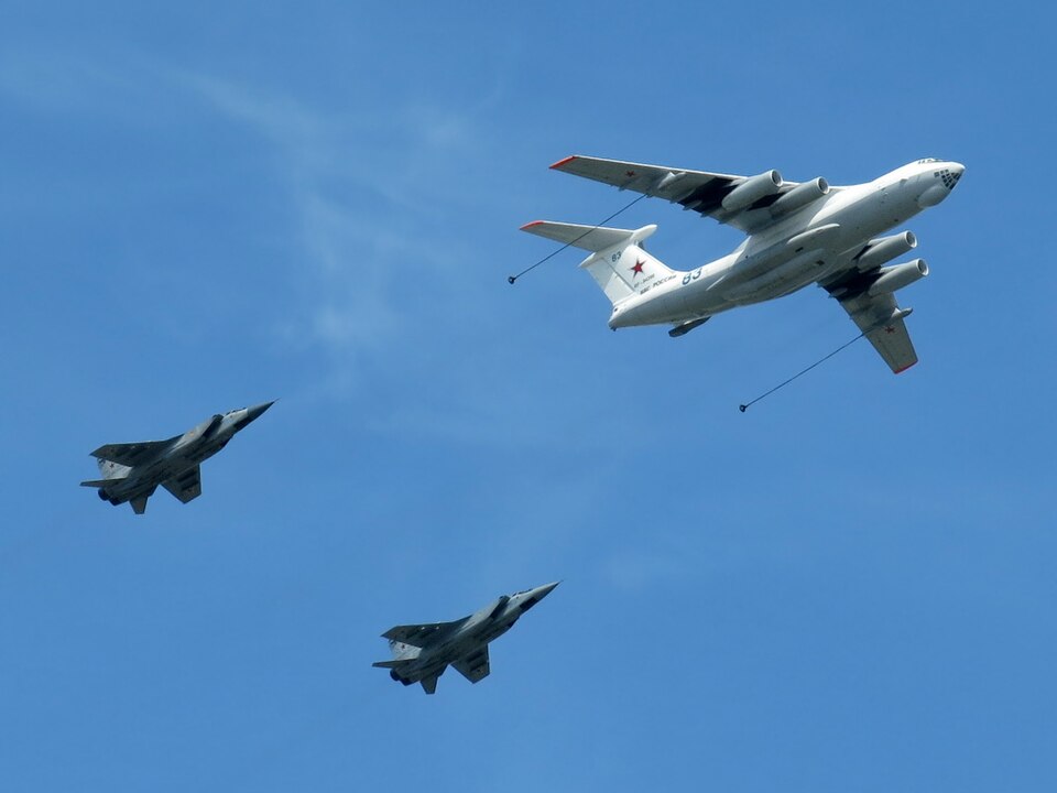 An Il-78M seen from below in flight, with its whole visible nose, four engine nacelles, wings, tail and extended refuelling hoses inside the frame; two MiG-31s fly behind it.