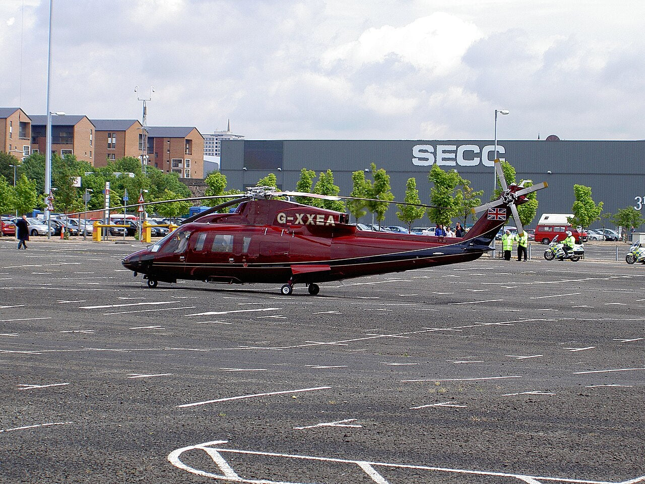 Burgundy S-76C parked in Glasgow, seen from the side with its cockpit, cabin windows, main rotor, tail rotor and wheels visible.