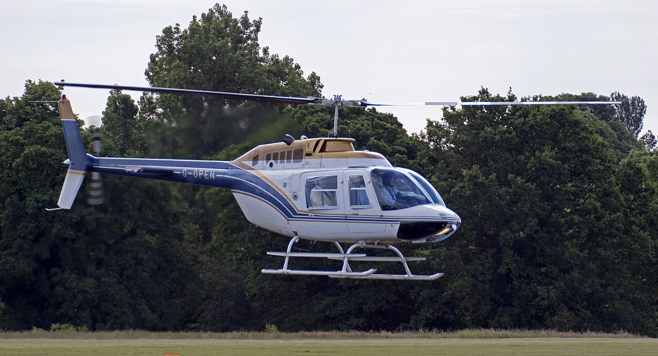 White, blue and gold Bell 206B G-OPEN hovering, with the complete visible main rotor, cabin, tail and skids inside the frame.