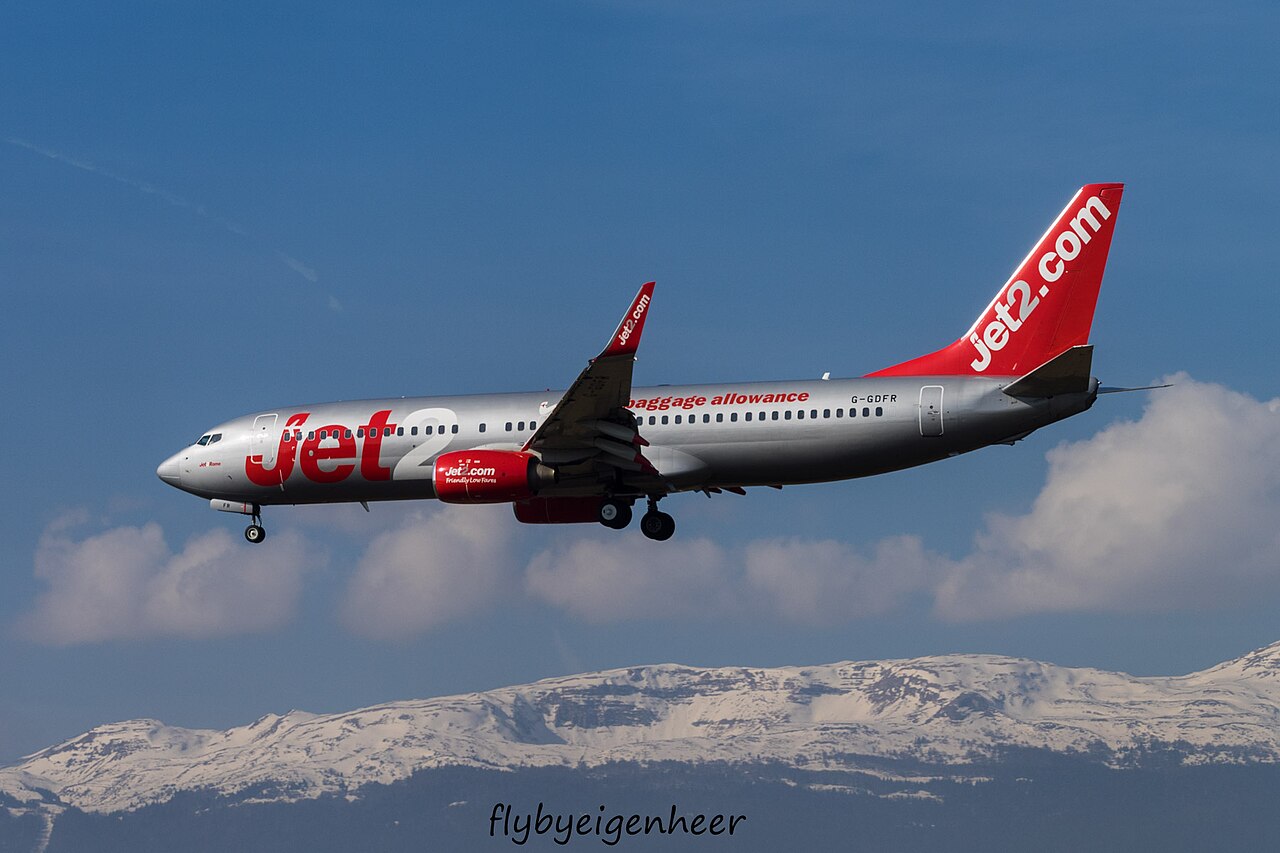 Full side and underside view of red-and-silver Boeing 737 G-GDFR in flight, with landing gear lowered, a raised wingtip and snowy mountains behind.