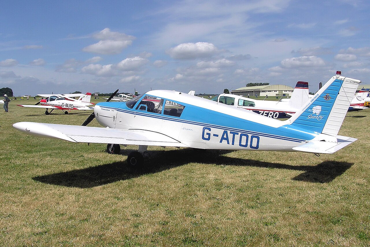 Blue-and-white Piper PA-28-140 Cherokee G-ATOO seen from the rear left on grass at Kemble, with its low wings, tail and wheels visible.