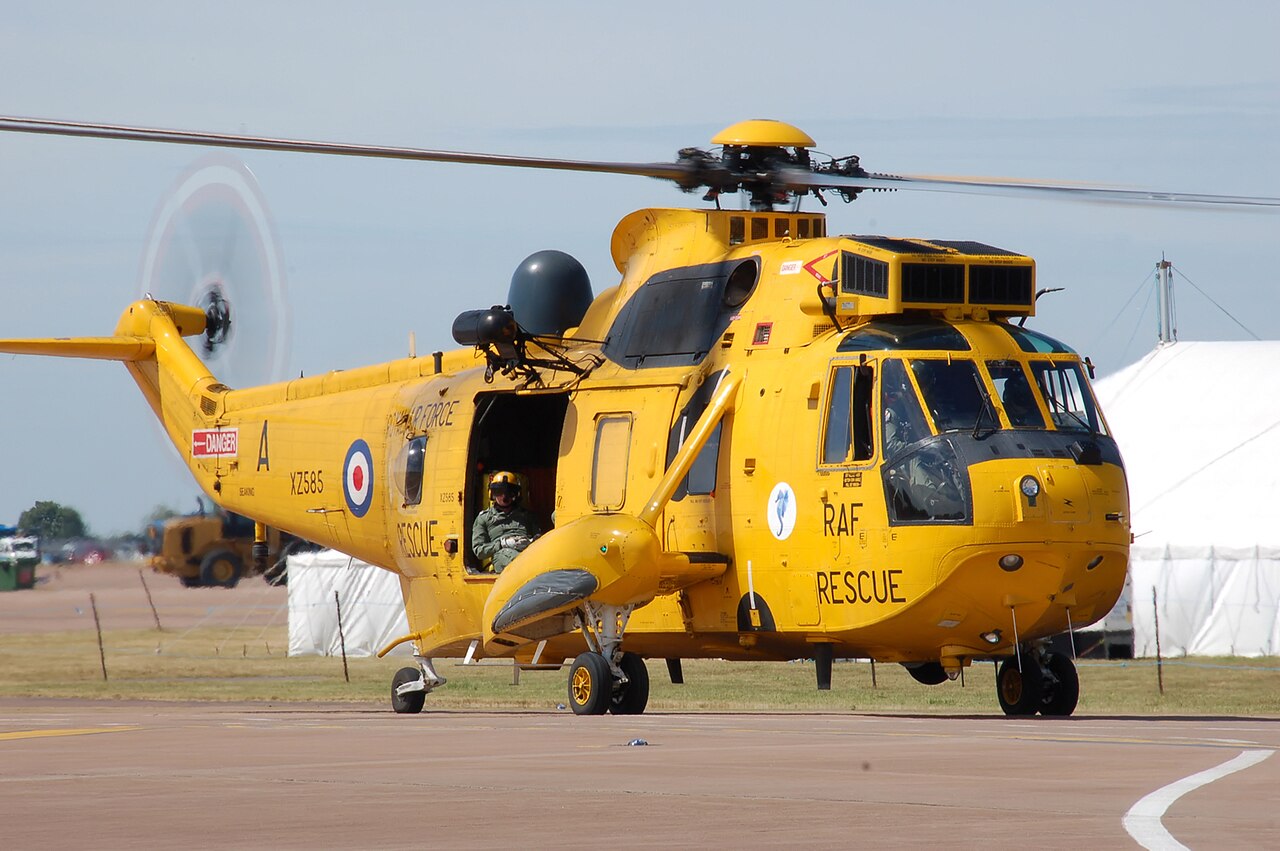 Yellow Sea King HAR3 viewed from the front and side, with an open side door and landing gear visible. Rotor tips extend beyond the frame.