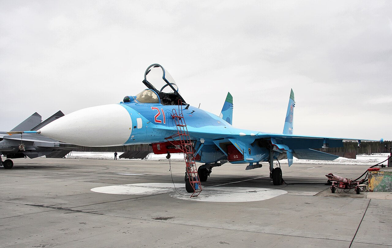 Blue and white Su-27 with one-seat cockpit, twin fins and broad wings, parked with its canopy open.