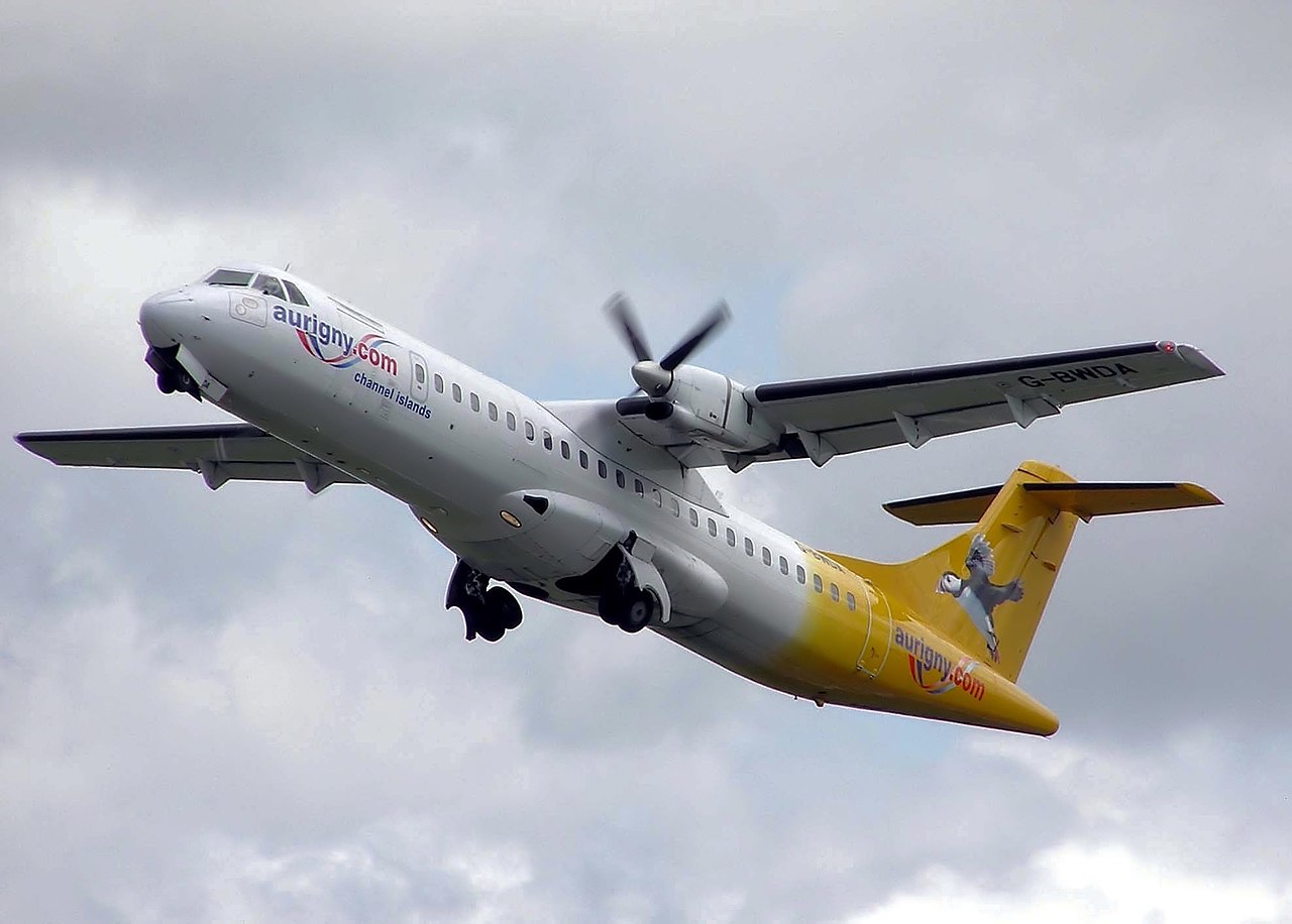White-and-yellow Aurigny ATR 72-202 G-BWDA climbing after takeoff, with a high wing, propeller blur and landing gear still visible.
