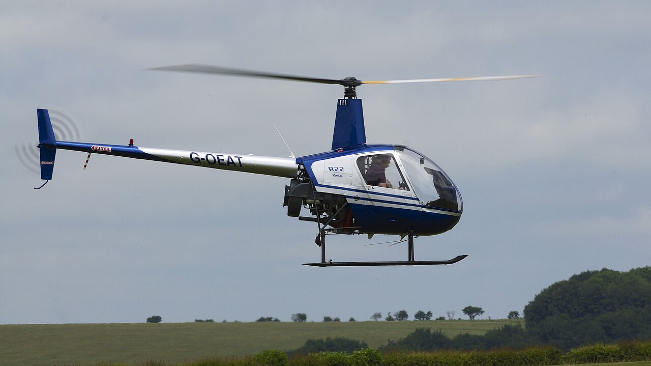 Blue-and-white Robinson R22 Beta G-OEAT hovering, with its entire visible rotor, cabin, tail boom and skids inside the frame.